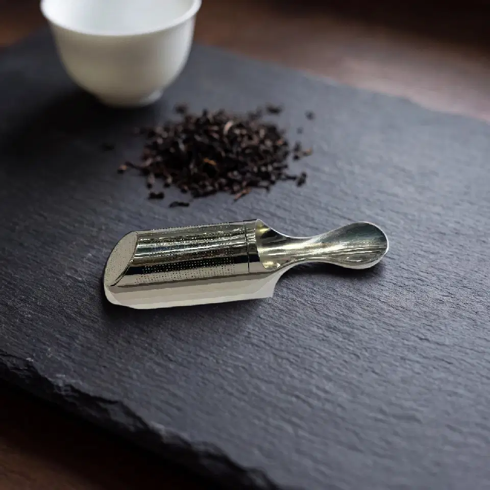 Un tube à thé en inox posé sur une ardoise, à côté de feuilles de thé noir et d'une tasse blanche