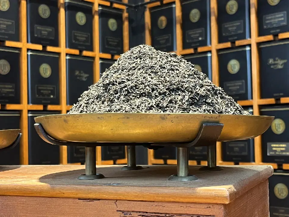 Feuilles de Smoky Earl Grey en vrac, en tas sur le plateau en laiton d'une balance, devant un mur de boîtes noires
