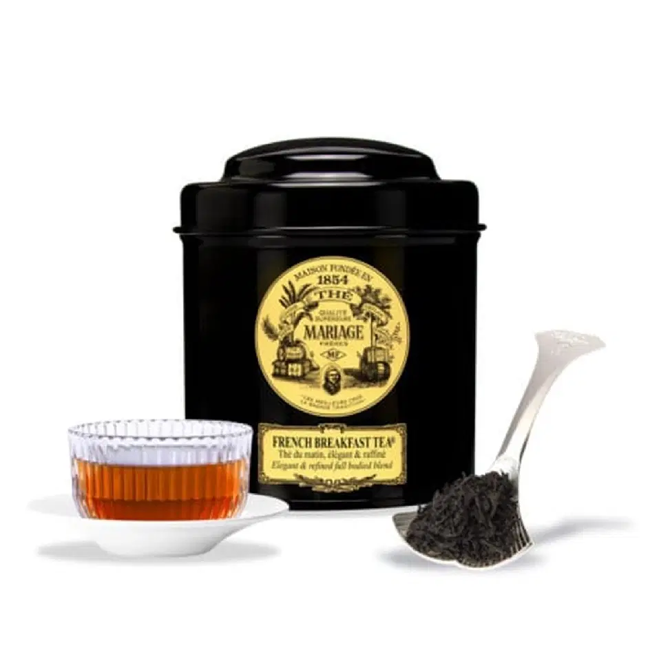 Boîte noire de French Breakfast Tea Mariage Frères à côté d'une tasse de thé ambré et d'une cuillère de feuilles